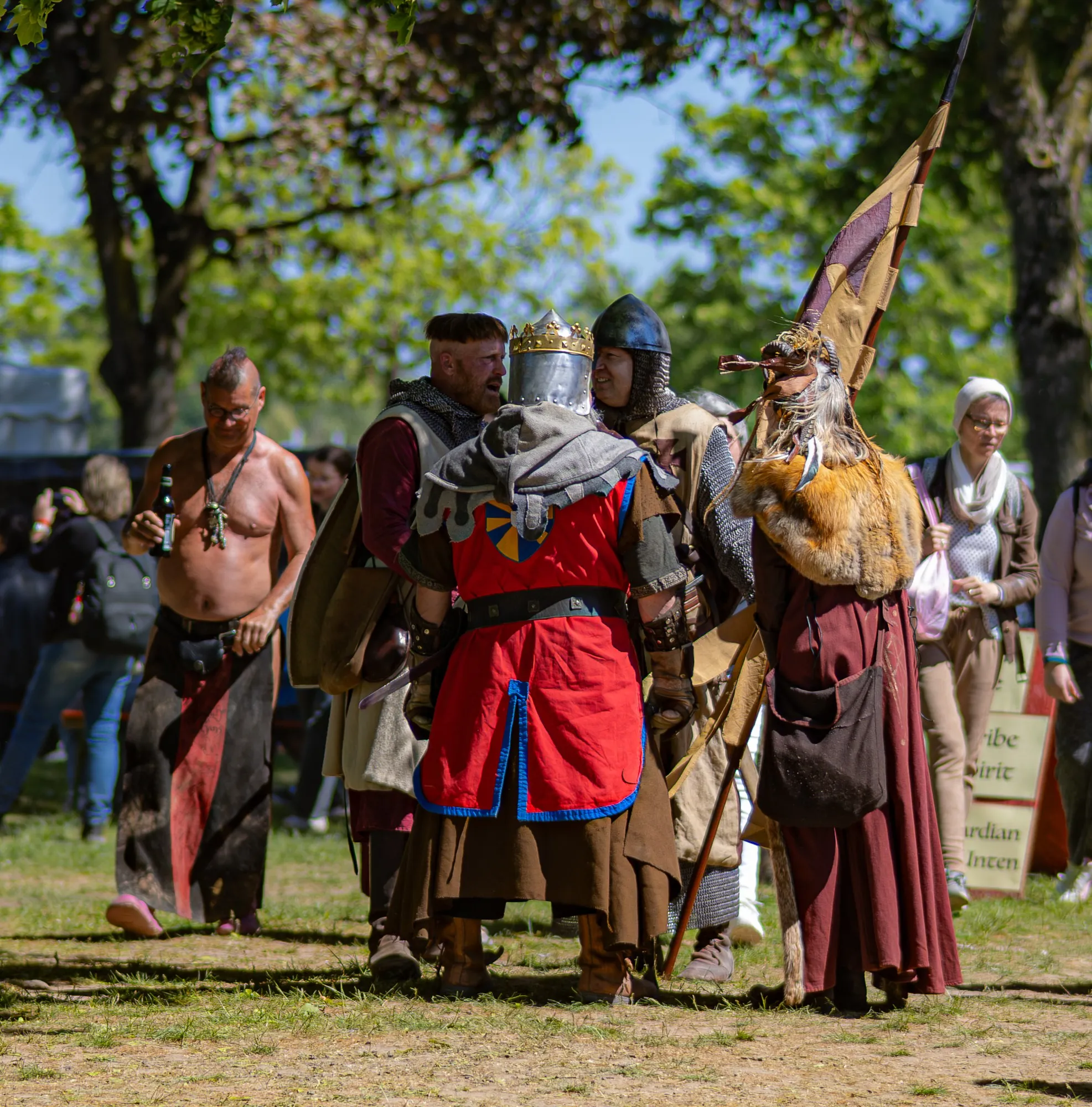 Gruppe von Reenactern auf einem Mittelaltermarkt im Gespräch – ein Ritter mit goldener Kronenhelm-Verzierung und rot-blauem Wappenrock, ein Kettenhemd-Kämpfer, eine mit Pelz und Federbanner ausgestattete Figur sowie ein barhäustiger Mann mit Bierflasche im Hintergrund.