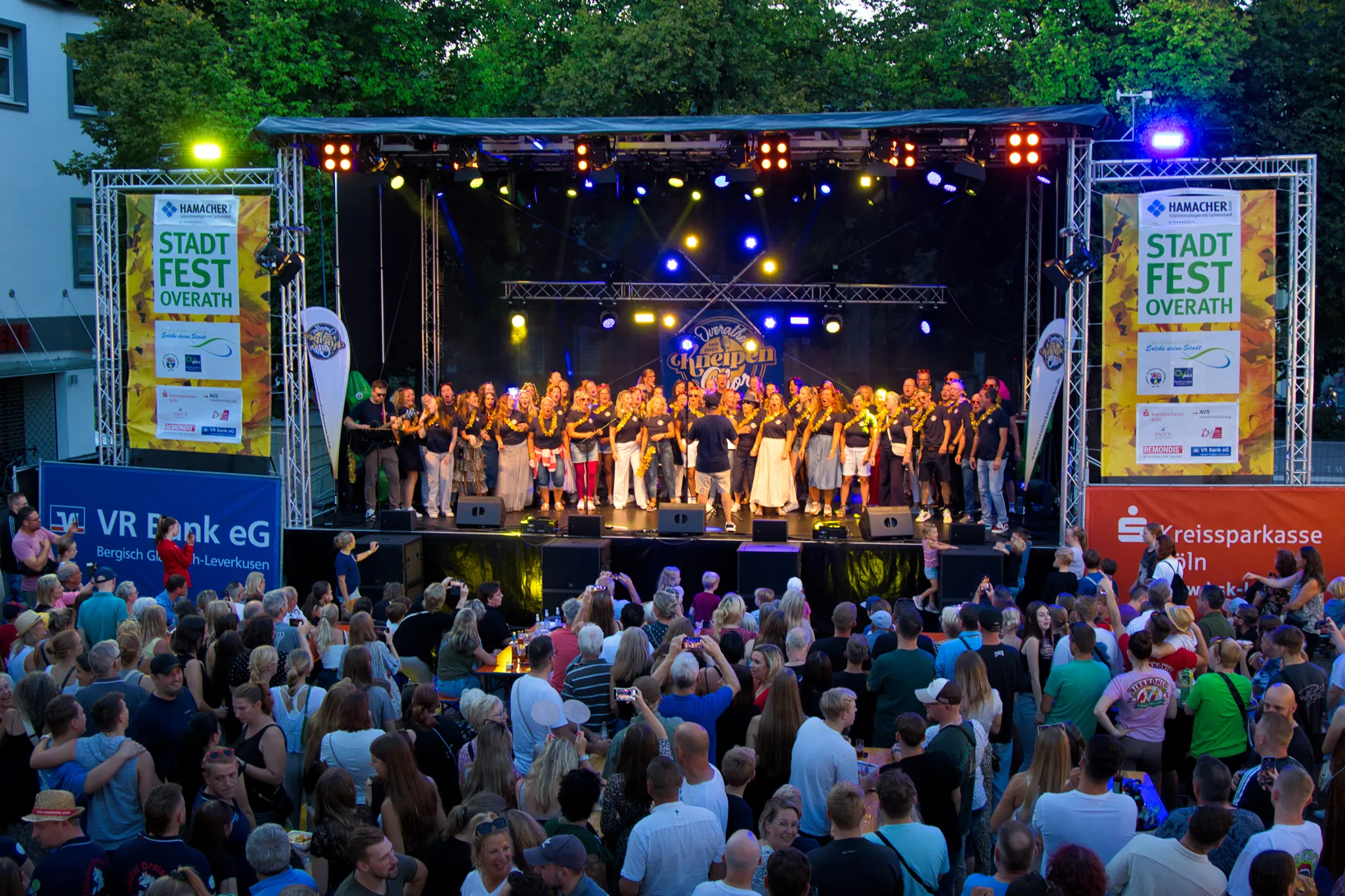 Weiter Blick von der Zuschauermenge aus auf die Bühne des Overather Kneipenchors bei einem Auftritt unter Bäumen, links und rechts Sponsorenbanner „Stadtfest Overath“.