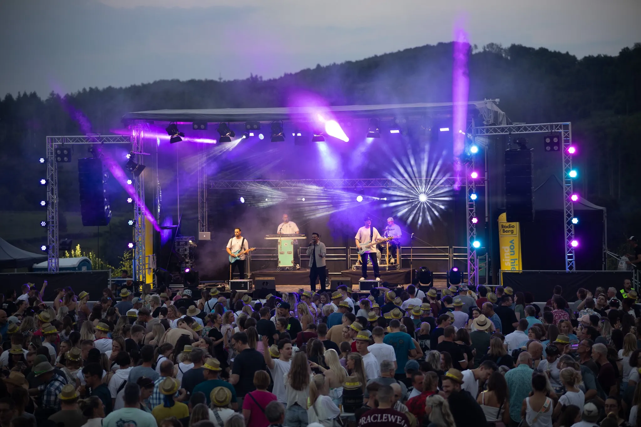 Weite Bühnenperspektive in der Abenddämmerung: Mehrere Musiker performen vor dichtem, jubelndem Publikum, im Hintergrund die Hügel des Bergischen Landes.