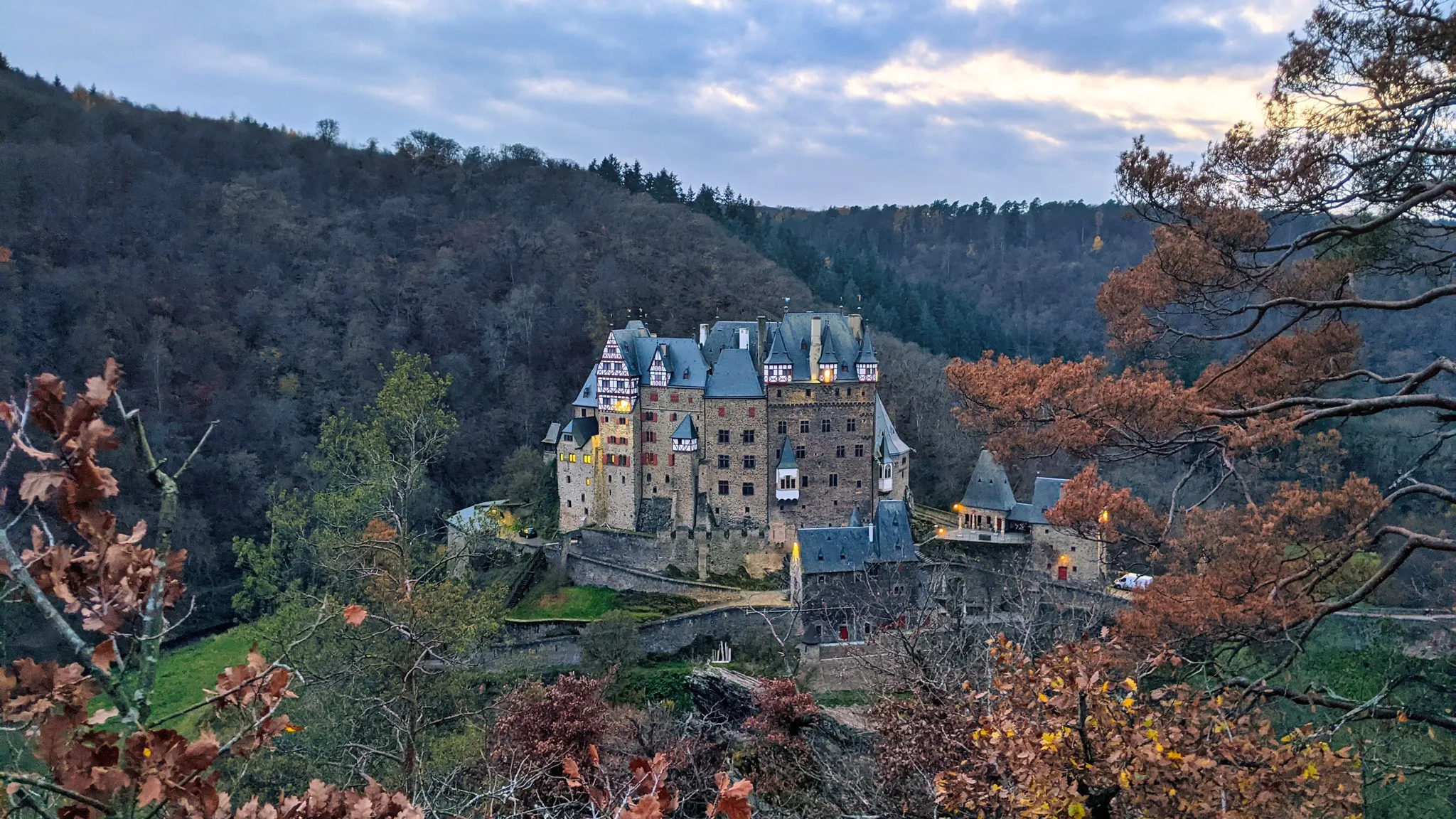 Die Burg Eltz thront in der blauen Stunde über dem herbstlich verfärbten Wald, ihre erleuchteten Fenster setzen einen warmen Kontrast zum kühlen Abendhimmel.