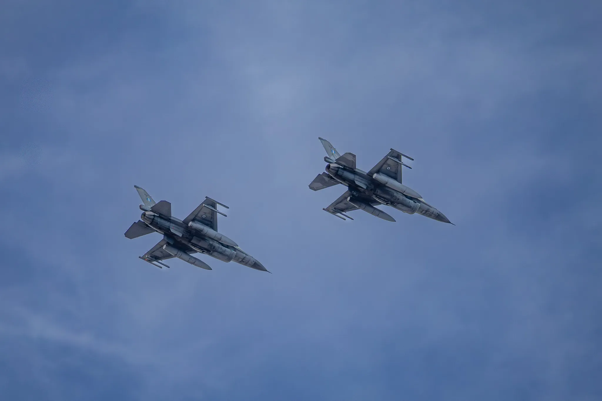 Zwei F-16 Fighting Falcon fliegen in enger Formation, von unten fotografiert vor leicht diesigem blauem Himmel.