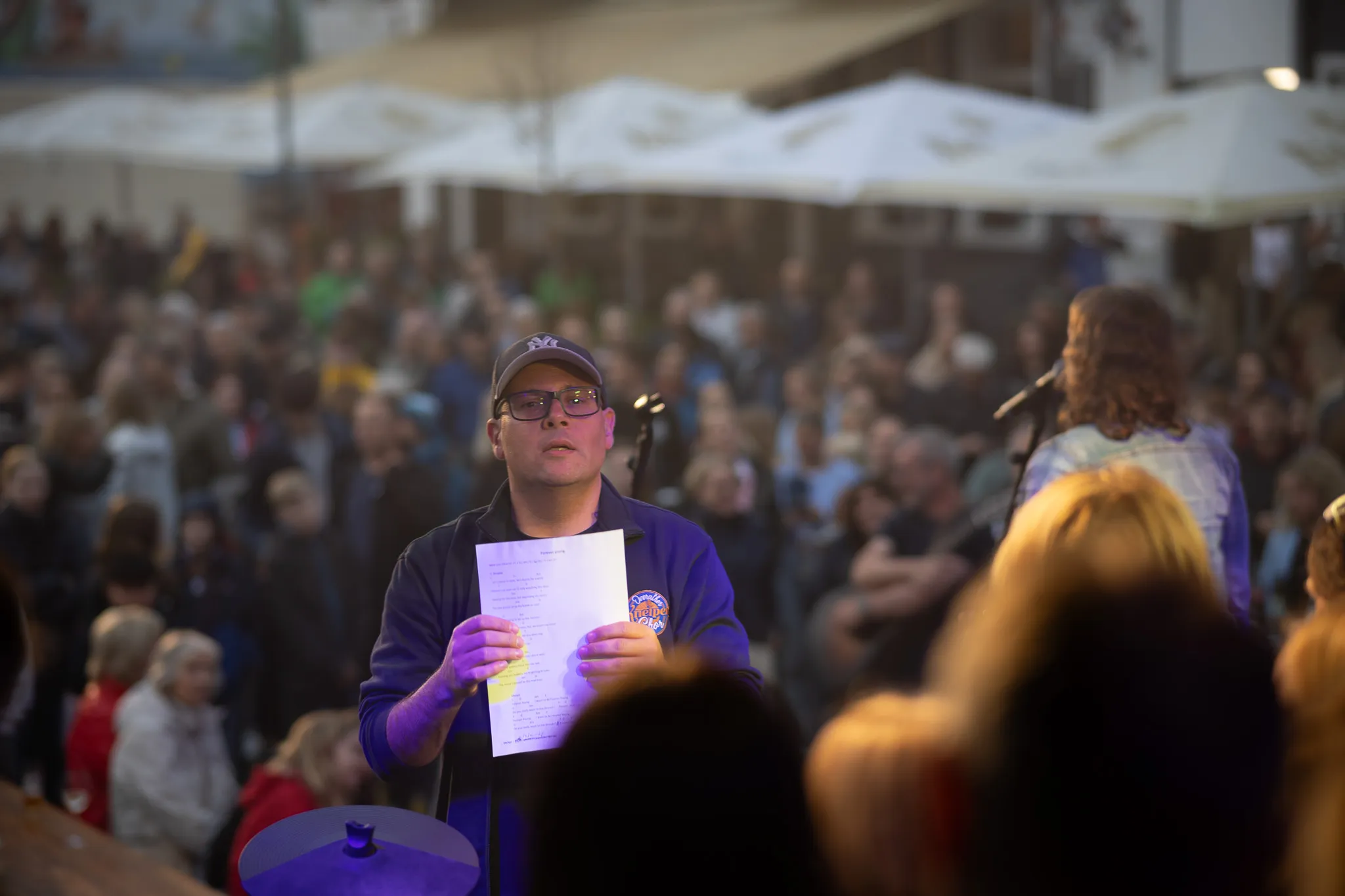 Ein Mann mit Baseballkappe und Brille hält ein Blatt mit Text in den Händen, während daneben eine weitere Person am Mikrofonständer zum Publikum gewandt steht, im unscharfen Hintergrund sitzt eine große Menschenmenge unter weißen Marktschirmen.