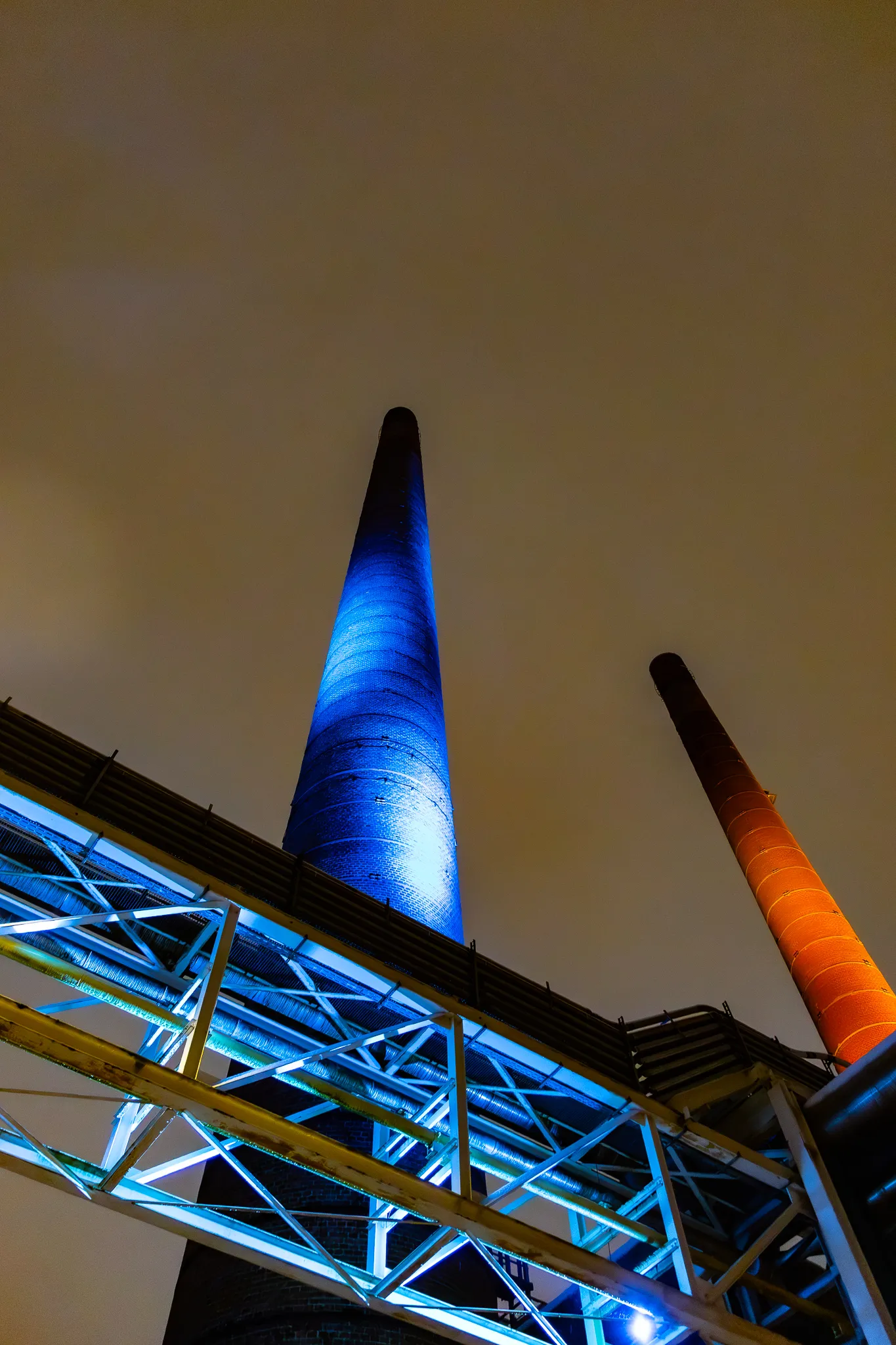Zwei hoch aufragende Schornsteine des Zanders Geländes, einer blau, einer orange angestrahlt, von unten fotografiert vor dunklem Nachthimmel.