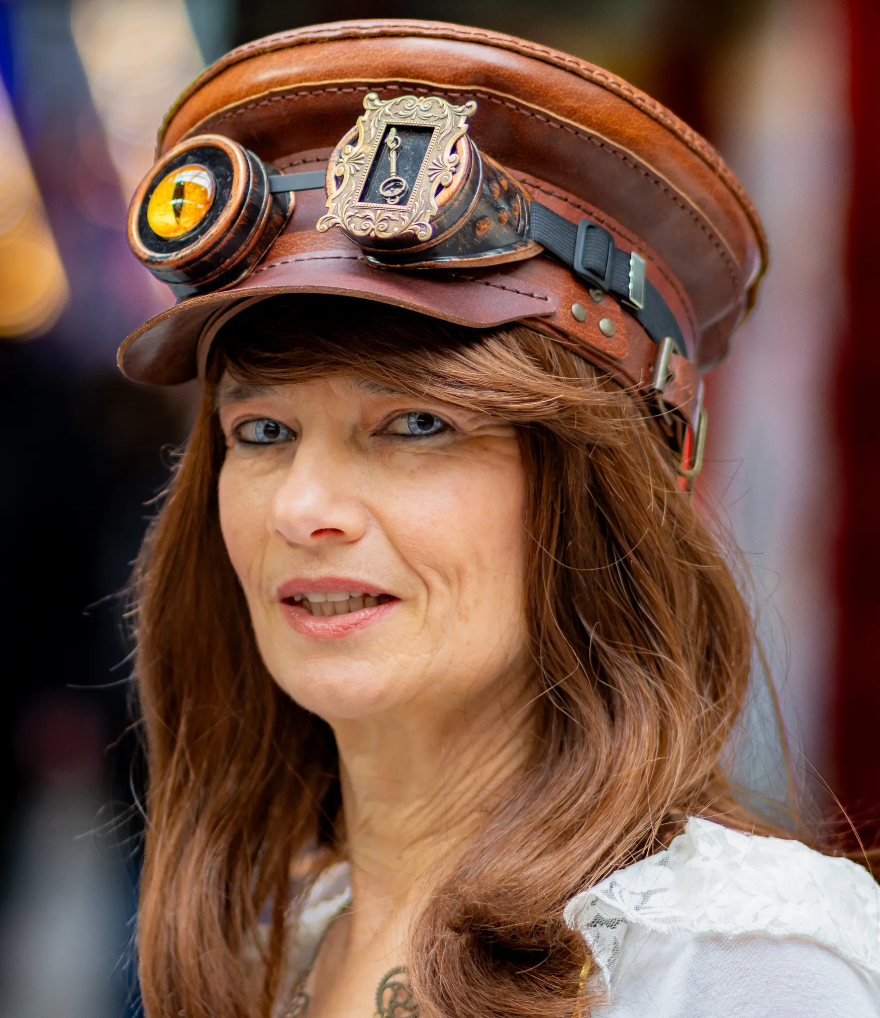 Frau mit braunem Lederkäppi und aufgesetzter Steampunk-Brille mit bernsteinfarbenem Glas, rotbraunes Haar, im Hintergrund unscharfe Jahrmarktlichter.