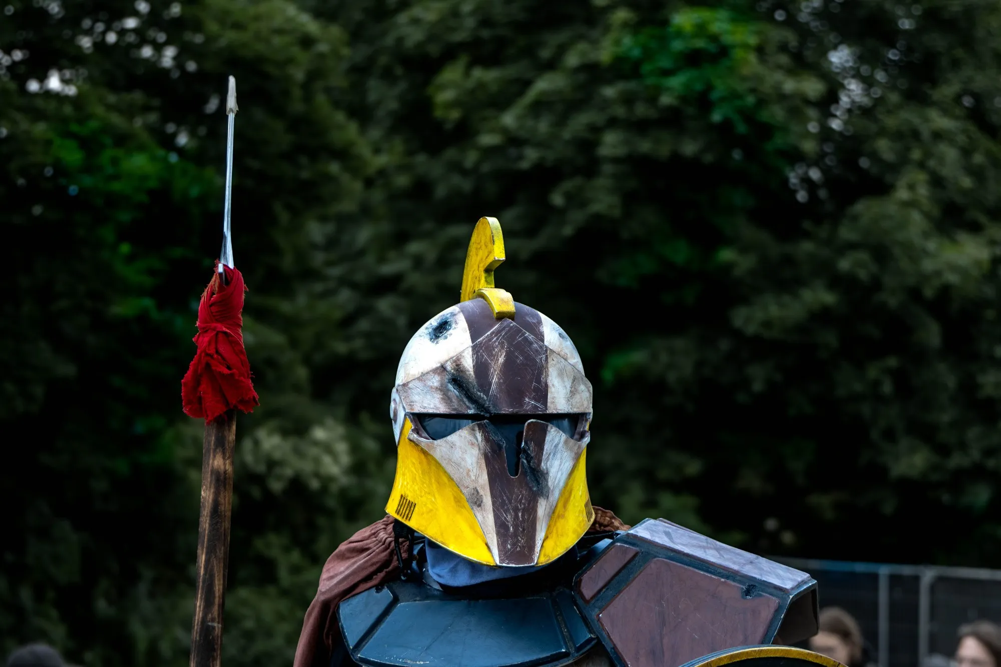 Ritter mit gelb-weiß gemustertem Helm und goldenem Federbusch, in dunkler Schulterrüstung, hält eine Lanze mit rotem Tuch vor bewaldetem Hintergrund.