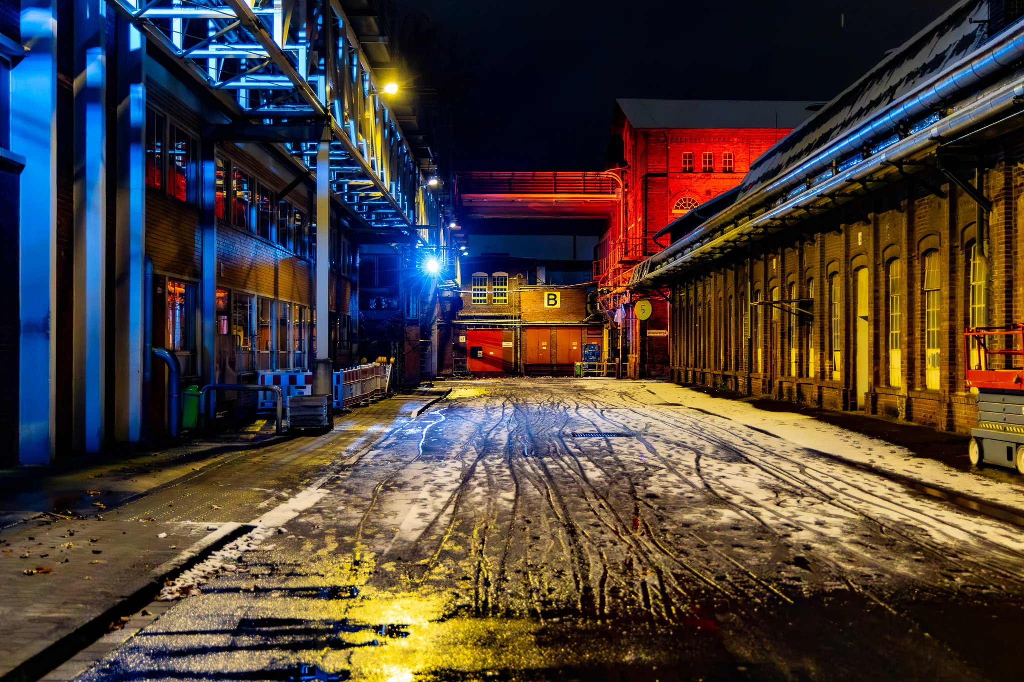 Blick durch eine Werksgasse auf dem Zanders Gelände: links blau illuminierte Stahlkonstruktion, rechts ein rot angestrahltes Backsteingebäude mit der Aufschrift „B“, der Boden ist schneebedeckt.