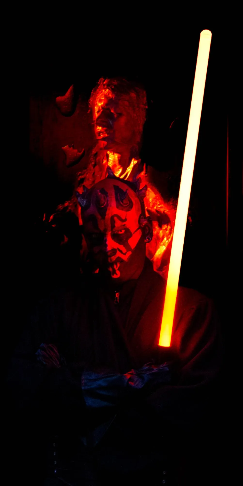 Cosplayer als geschminkter Sith-Charakter mit schwarzer Robe, rot-schwarzem Gesichtstattoo und Hörnern, hält ein leuchtendes Lichtschwert vor dunklem, rötlich beleuchtetem Hintergrund.