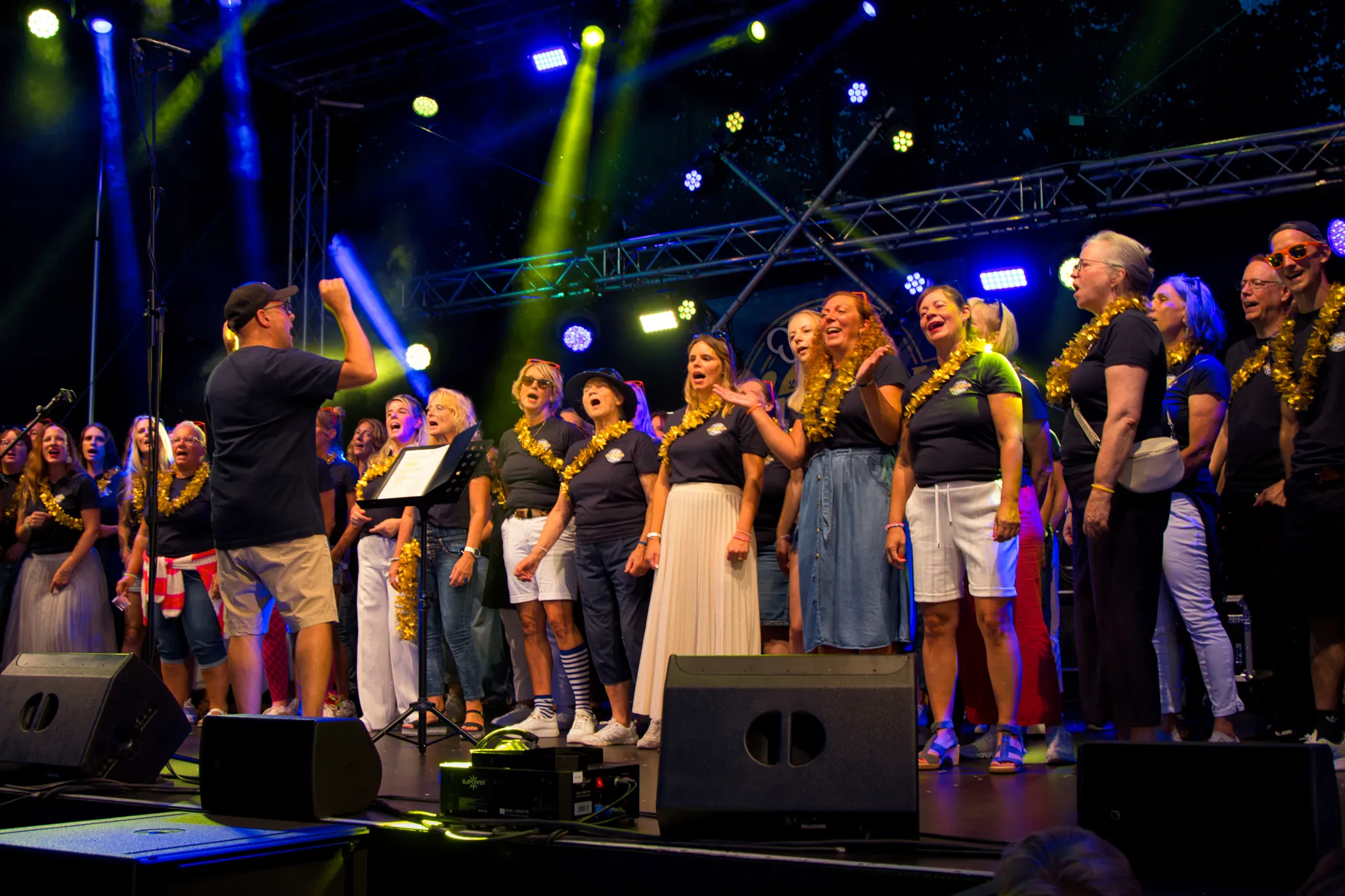 Chorleiter dirigiert von der Bühne aus die applaudierenden und singenden Chormitglieder, im Hintergrund grün-blaues Bühnenlicht.