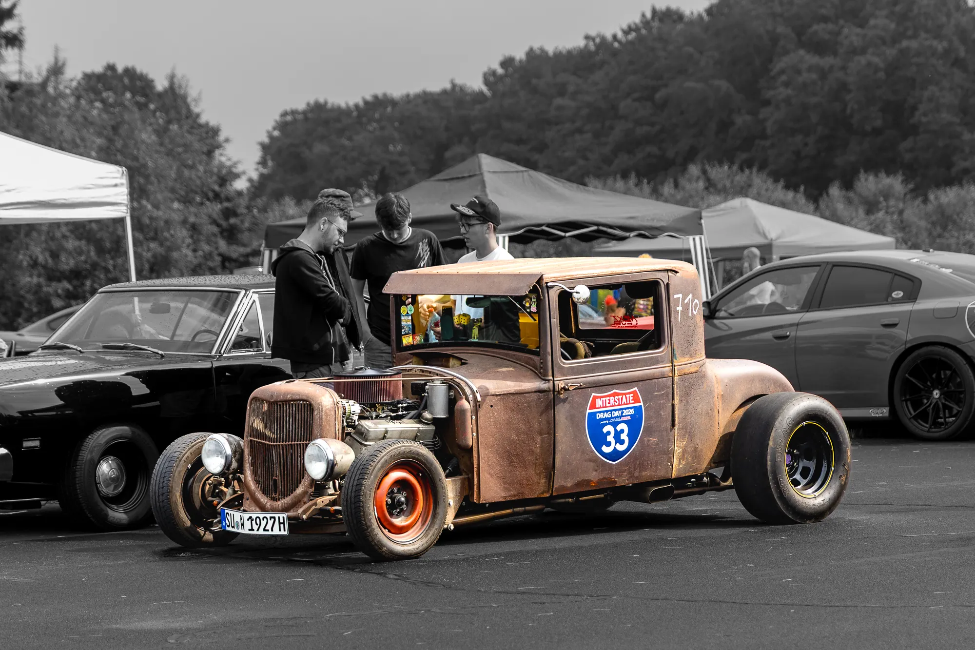 Rostiger Ford Hot-Rod mit offener Motorhaube, freiliegendem Motor und Interstate-Drag-Day-2026-Aufkleber, drei Besucher unterhalten sich am Fahrzeug, im Hintergrund Pavillons und Bäume.