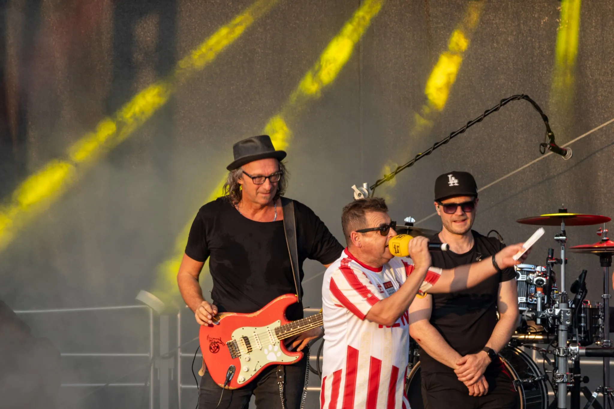 Dreiköpfige Band auf der Bühne: Ein Gitarrist mit Hut und roter E-Gitarre spielt neben einem Sänger im gestreiften Trikot, der ins gelbe Mikrofon singt.