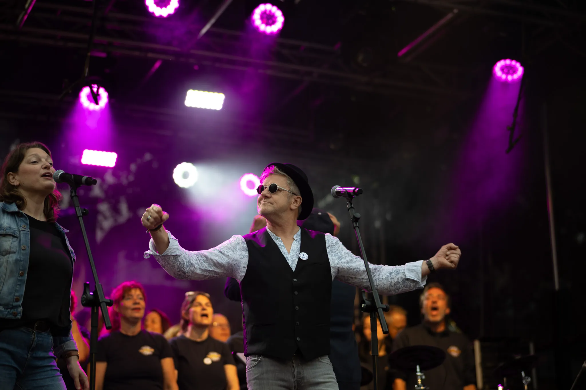 Eine Sängerin in blauer Jeansjacke singt konzentriert in ein Standmikrofon, während daneben ein Mann mit Sonnenbrille und schwarzer Mütze mit weit ausgebreiteten Armen und geballten Fäusten eine triumphierende Geste macht, beide stehen im grellen Bühnenlicht.
