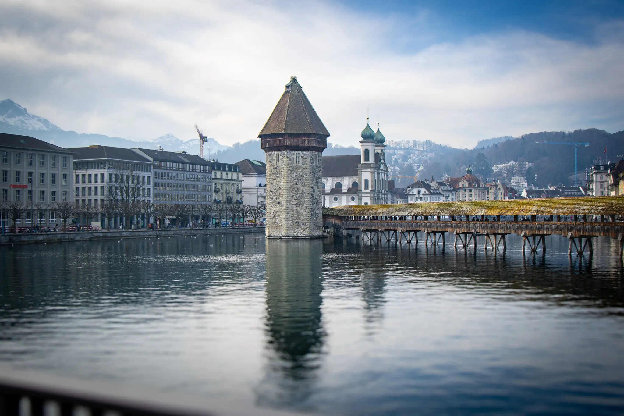 Der Kapellbrückenturm mit der historischen Kapellbrücke über der Reuss in Luzern, im Hintergrund die Kirche St. Peter und schneebedeckte Alpengipfel im weichen, diffusen Licht.