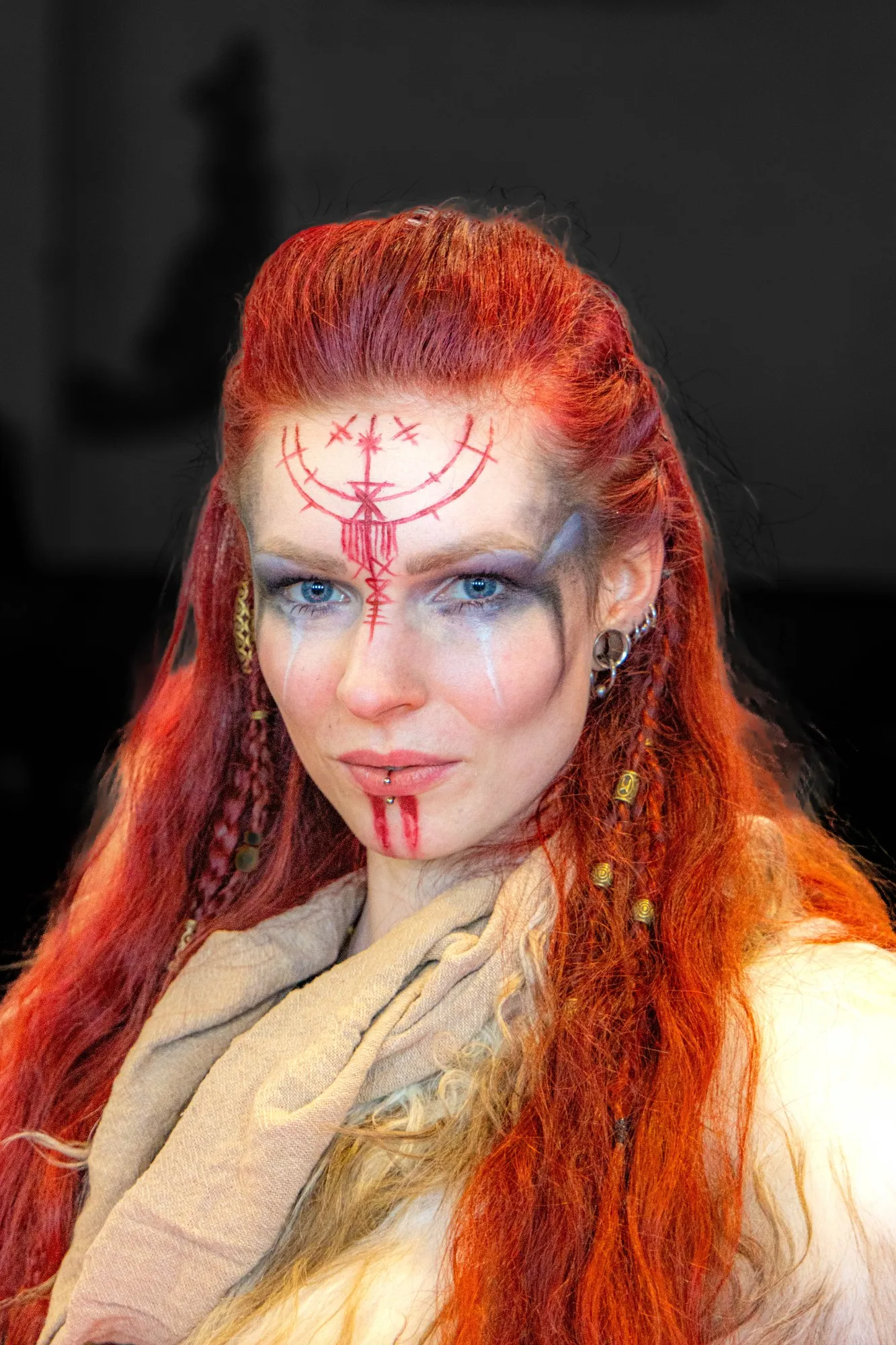 Porträt einer Fantasy-Kriegerin mit rot geschminkten Runen auf der Stirn, dunkel umrandeten Augen und braunem Tuch um den Hals vor dunkel verschwommenem Hintergrund.