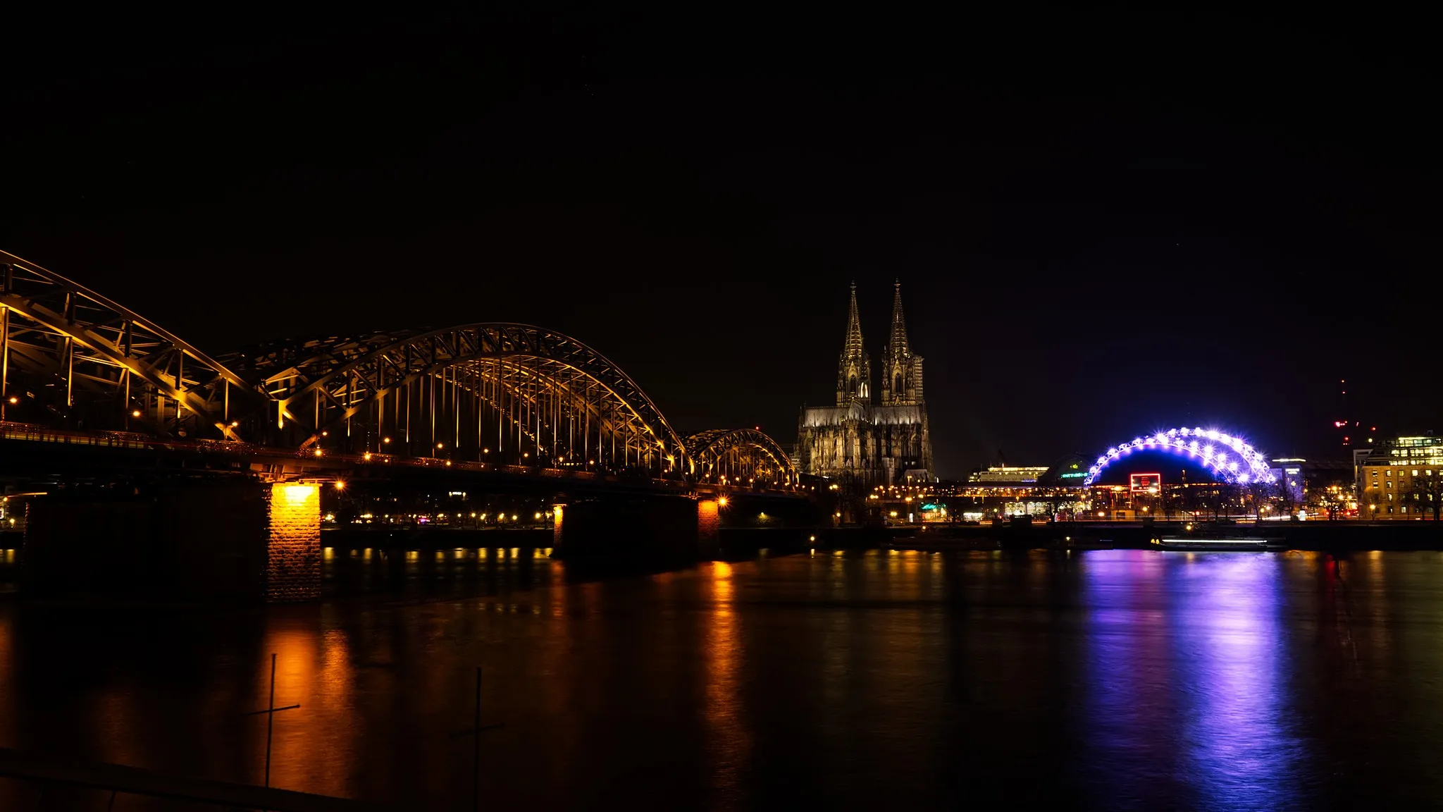 Nächtliche Skyline von Köln mit beleuchteter Hohenzollernbrücke, dem Kölner Dom und dem blau illuminierten Rheinkran, gespiegelt im Rhein.