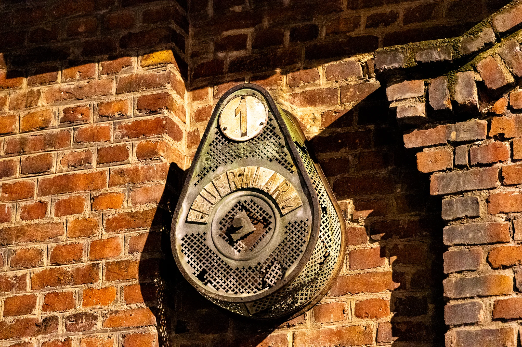 Historisches Feueralarm-Schild aus Metall an einer Backsteinwand des Zanders Geländes, warm angeleuchtet, mit der Nummer 1 und durchbrochenem Lochmuster.