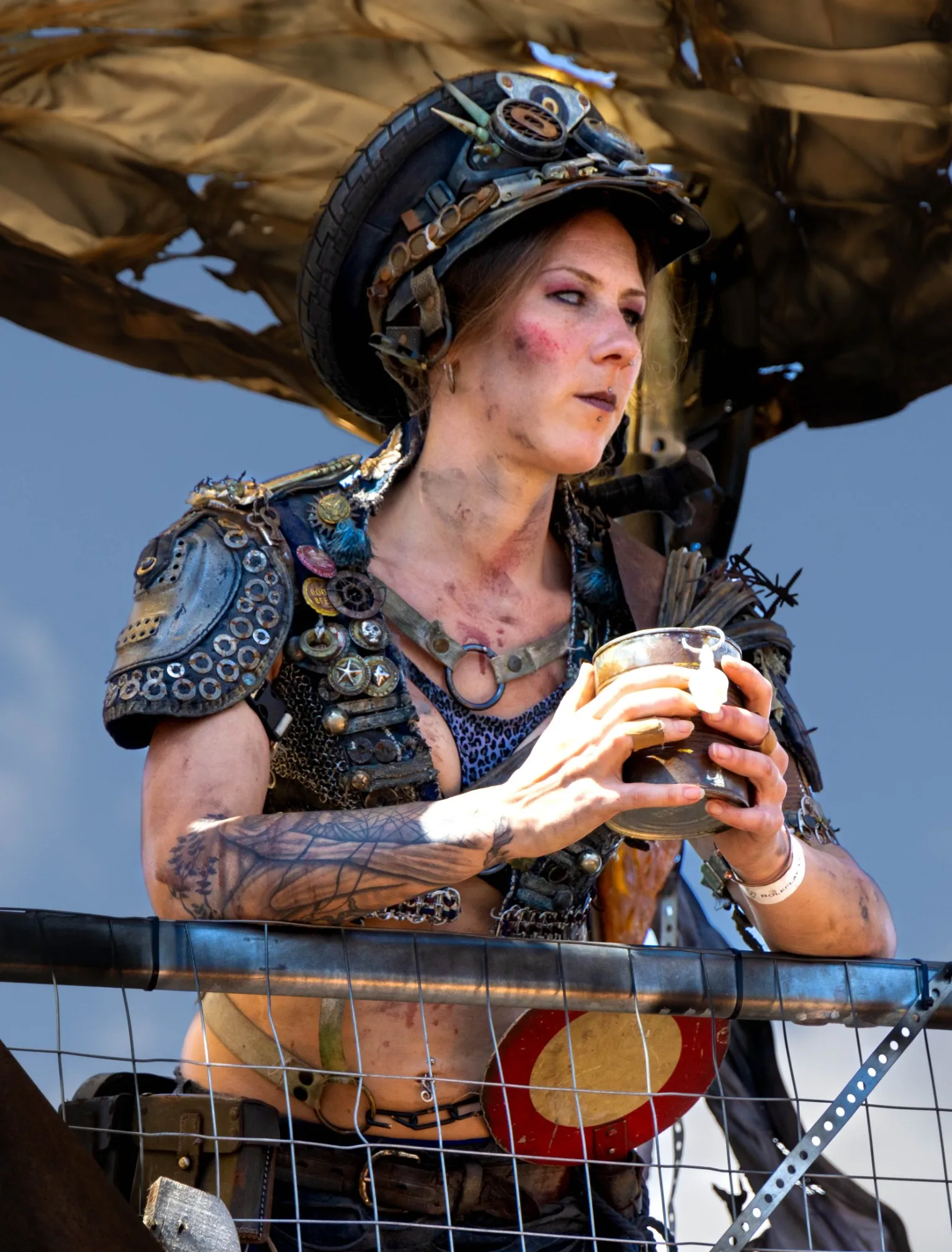 Cosplayerin in Endzeit-Kostüm mit Autoreifen-Helm, Schulterpanzer aus Metallscheiben und Leopardenmuster-Top, lehnt an einem Metallgitter und hält einen Becher, Blick zur Seite.
