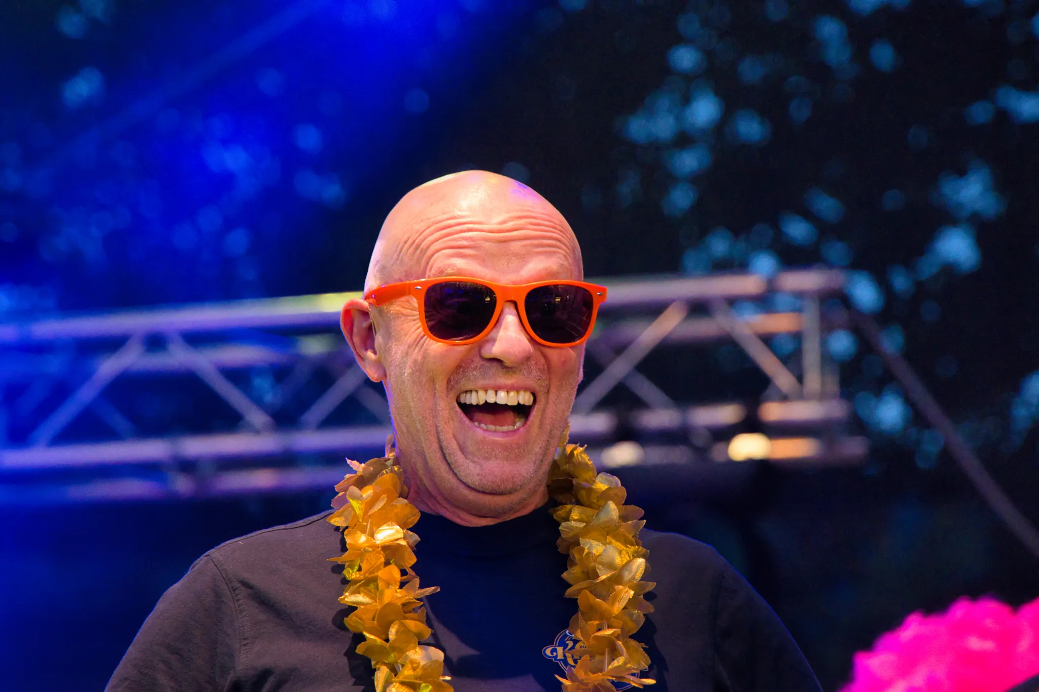 Nahporträt eines lachenden Chormitglieds mit Glatze, orangefarbener Sonnenbrille auf dem Kopf und goldener Blumenkette.