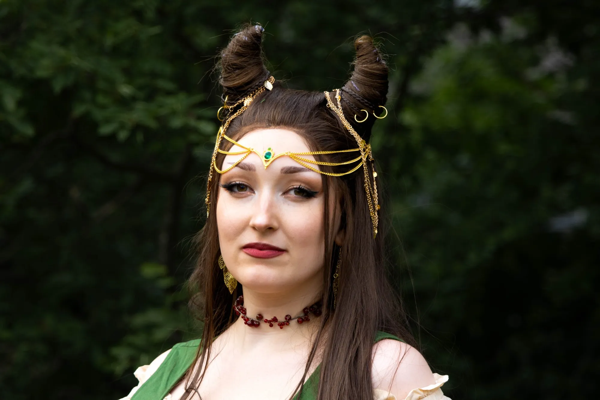 Porträt einer Cosplayerin mit geflochtenen, hornförmigen Haarteilen, goldenem Kettenkopfschmuck mit grünem Stein, roter Beerenkette und grünem Kleid vor dunklem Waldhintergrund.