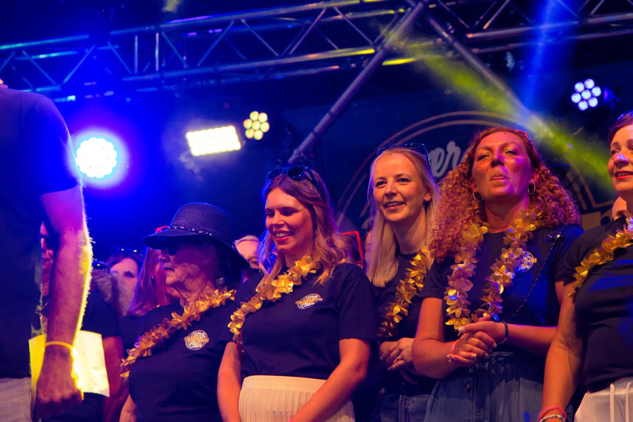 Gruppe lächelnder Sängerinnen mit goldenen Blumenketten dicht beieinander auf der Bühne, eine Frau mit Hut und Sonnenbrille im Vordergrund.