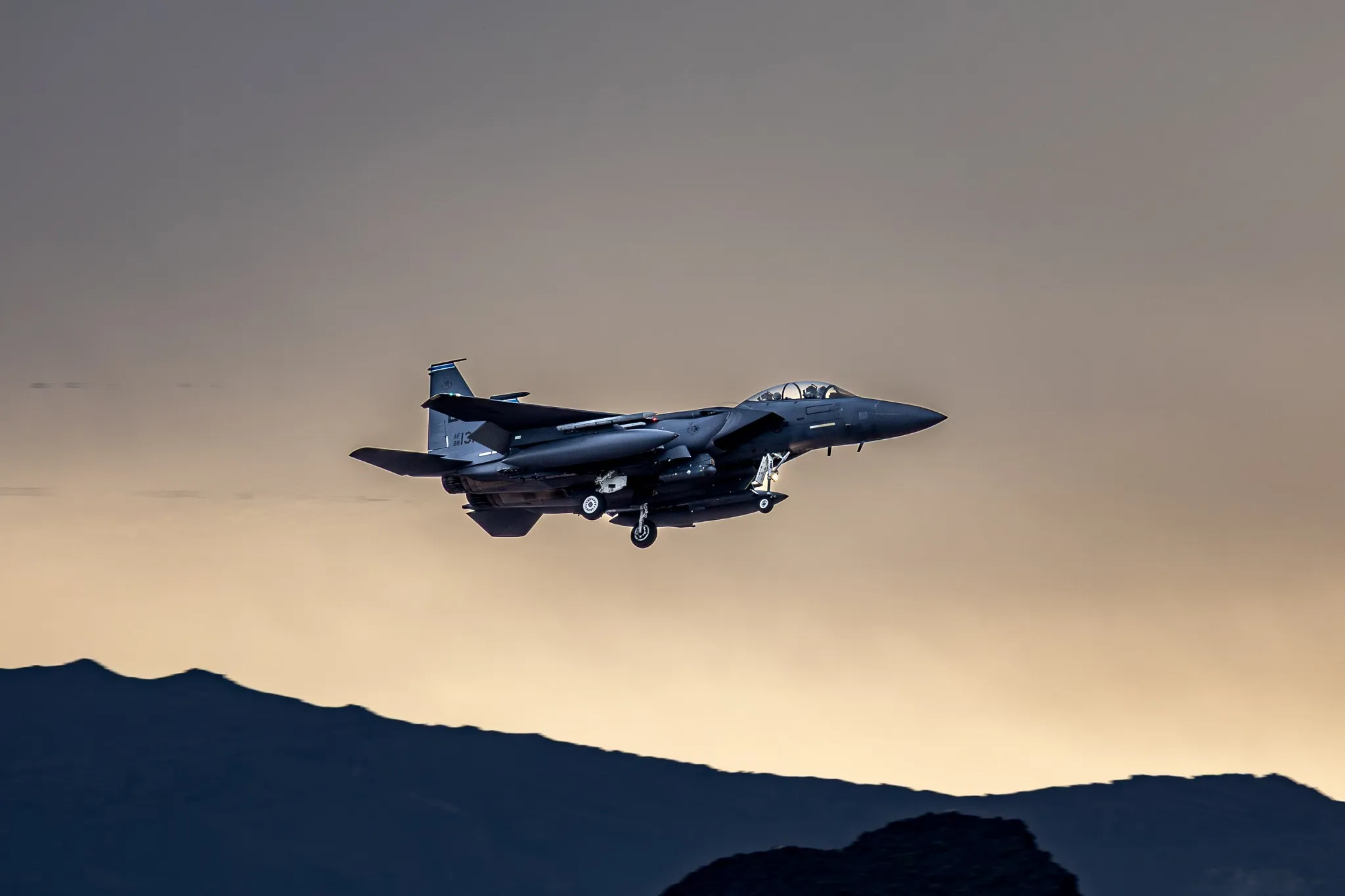 Eine F-15E Strike Eagle im Landeanflug mit ausgefahrenem Fahrwerk vor einer Bergsilhouette im goldenen Abendlicht.