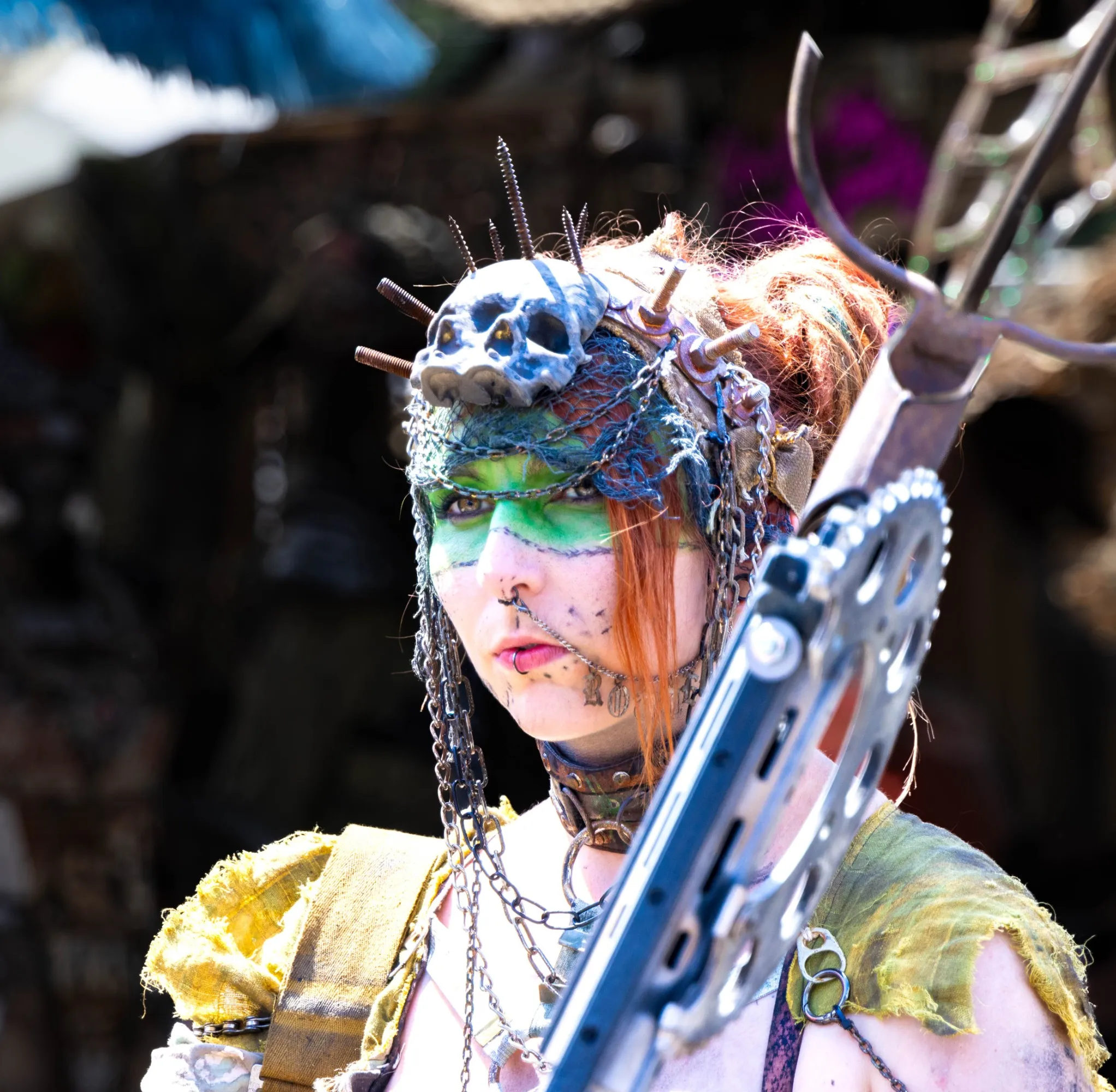 Nahporträt einer Endzeit-Kriegerin mit Tierschädel-Kopfschmuck, Metallspikes im Haar, grüner Tribal-Gesichtsbemalung und Kettenschmuck, hält eine gezackte Metallwaffe vor der Kamera.