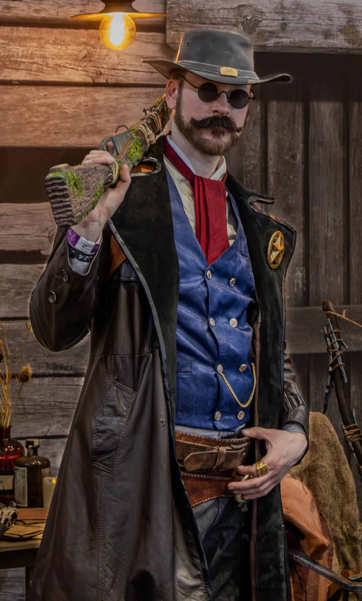 Cosplayer im Westernstil mit grauem Lederhut, runder Sonnenbrille, Vollbart und Sheriffstern auf schwarzem Ledermantel, hält ein bemoostes Gewehr über der Schulter vor rustikaler Holzwand.