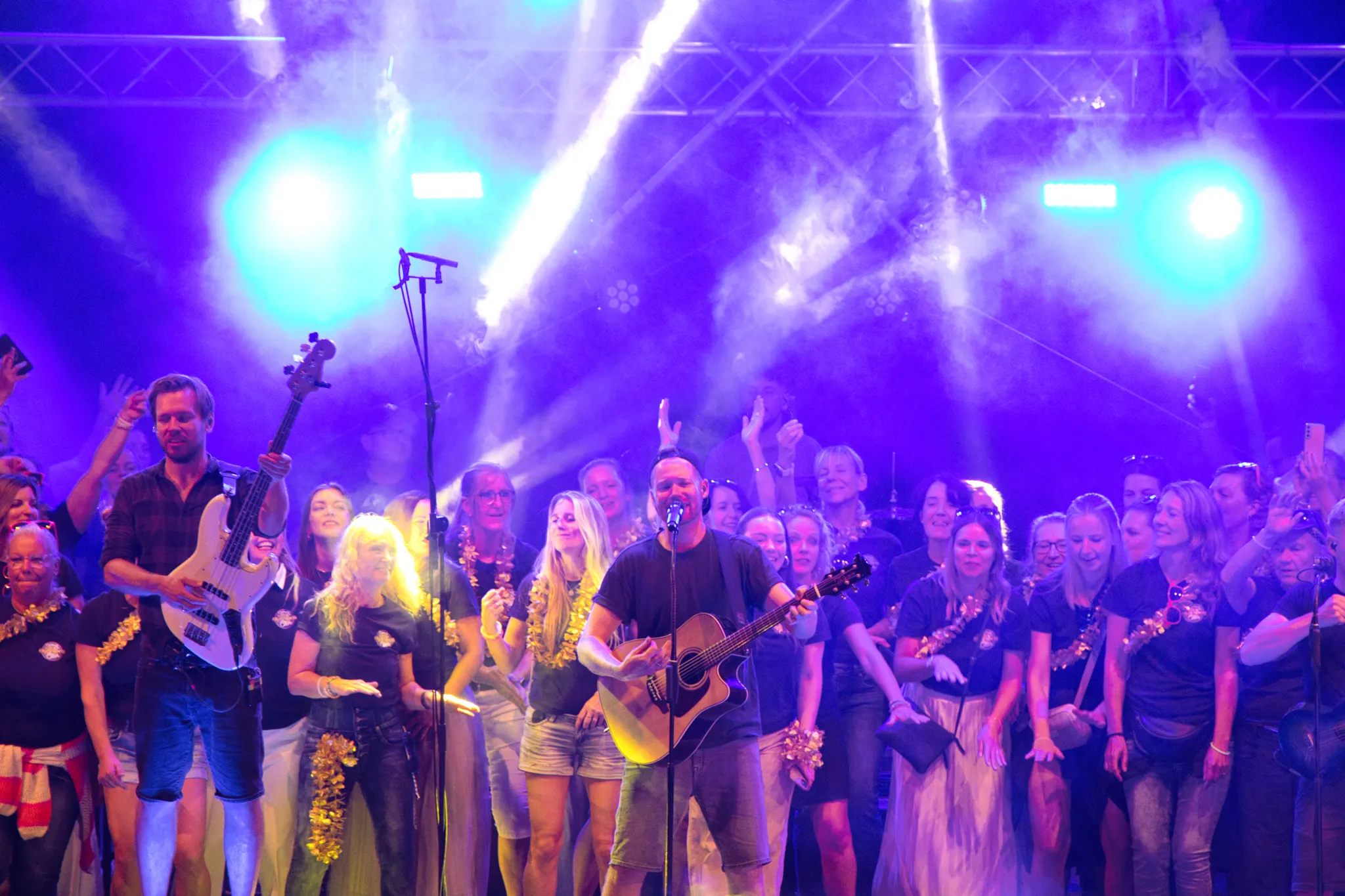 Bassist und Gitarrist der Begleitband inmitten des Chors, im Vordergrund zwei Chormitglieder in herzlicher Umarmung, warmes Bühnenlicht.