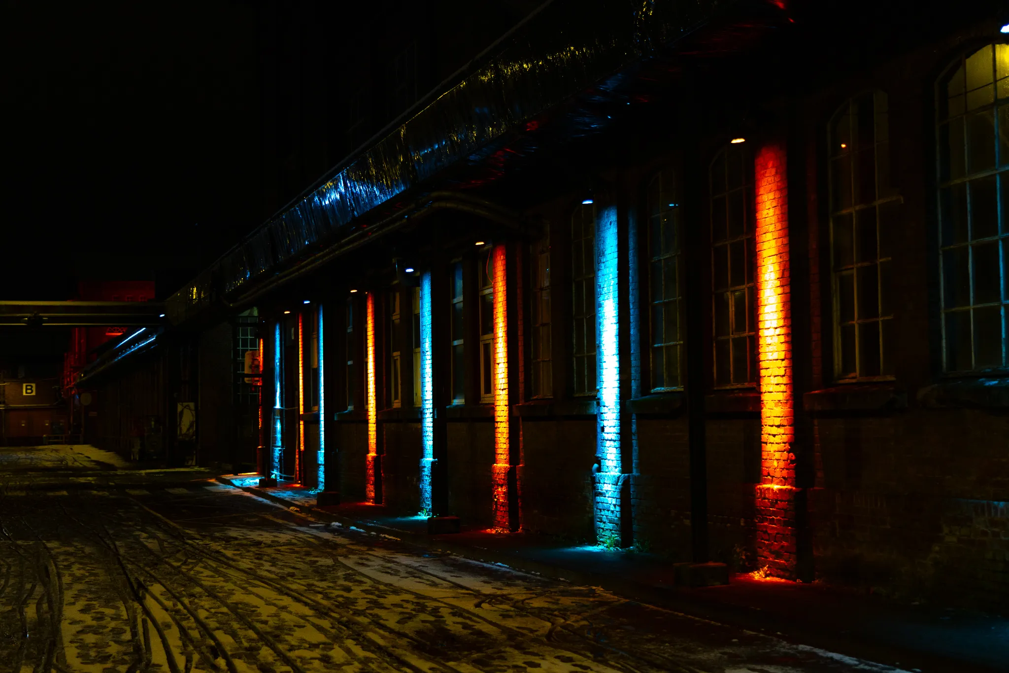 Reihe historischer Backsteinpfeiler, abwechselnd in Blau, Orange und Rot angestrahlt, mit Sprossenfenstern dazwischen – die Lichtinstallation auf dem Zanders Gelände bei Nacht.