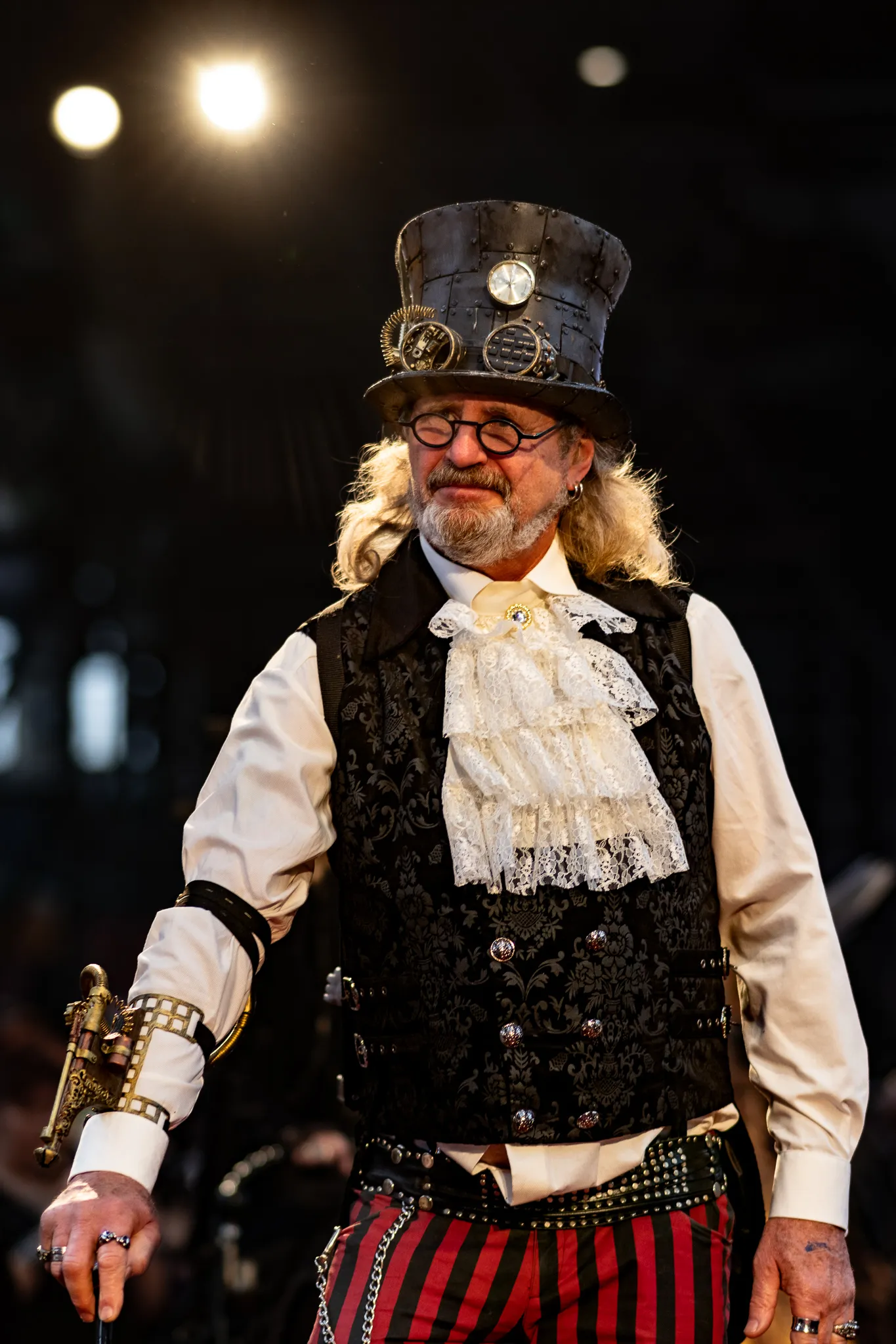 Mann mit runder Brille, grauem Bart und genietetem Steampunk-Zylinder mit Zahnrad-Applikationen, weißem Rüschenjabot und rot-schwarz gestreifter Hose, hält eine dekorative Pistolen-Requisite.