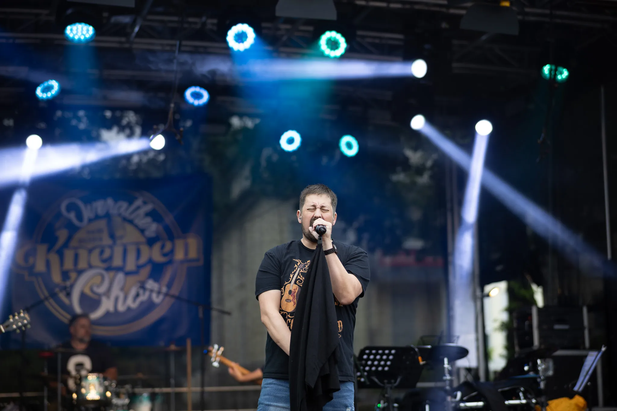 Ein Sänger mit Bart und schwarzem T-Shirt mit orangefarbenem Gitarrenmotiv singt mit geschlossenen Augen nah am Mikrofon, im Hintergrund ist unscharf ein Schlagzeuger zu erkennen und ein Banner mit der Aufschrift „Overather Kneipenchor” sichtbar.