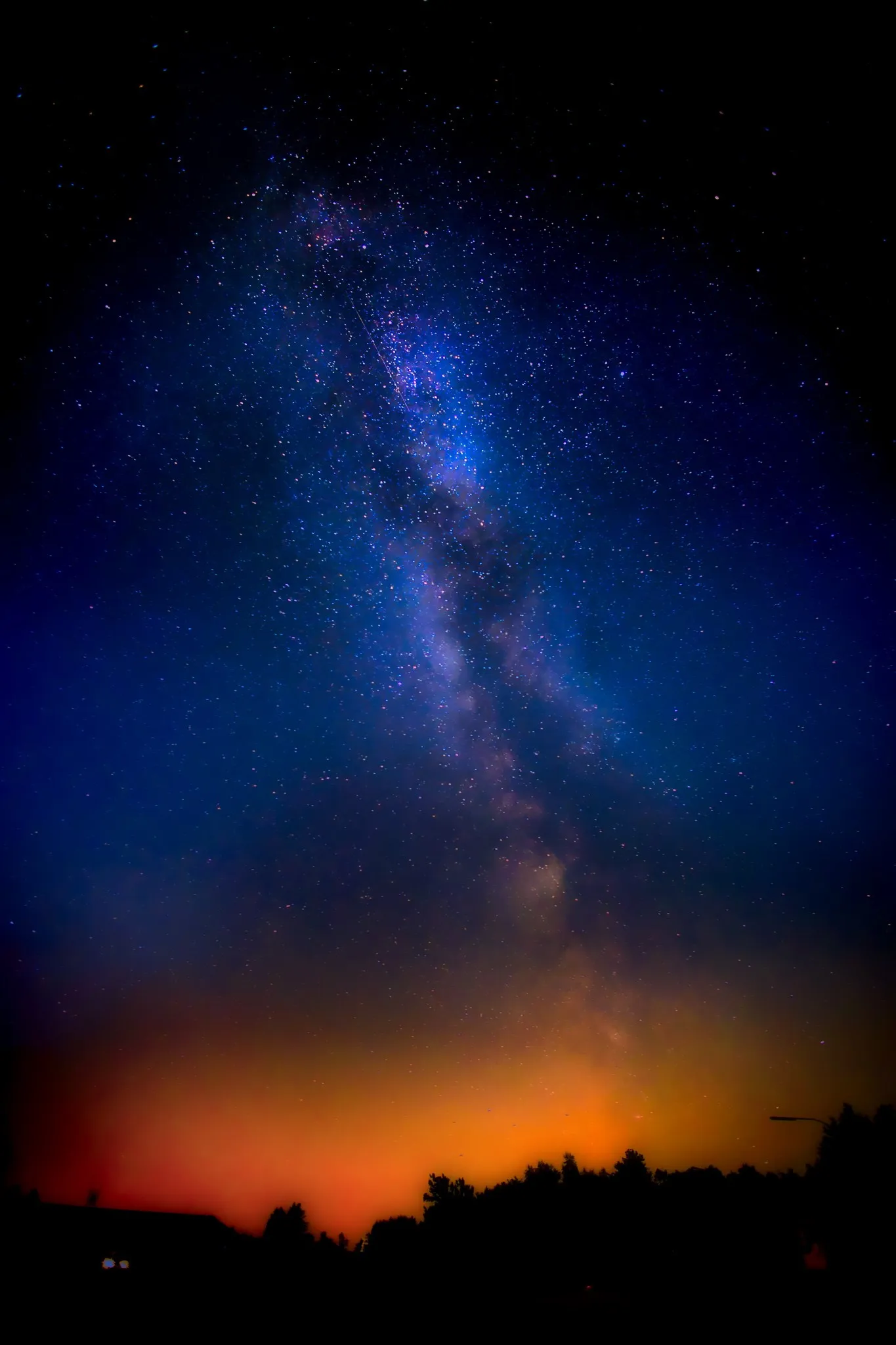 Milchstraßenband zieht sich als heller, sternenreicher Streifen über einen tiefblauen Nachthimmel bis zu einem orange erleuchteten Horizont mit dunkler Baumsilhouette.