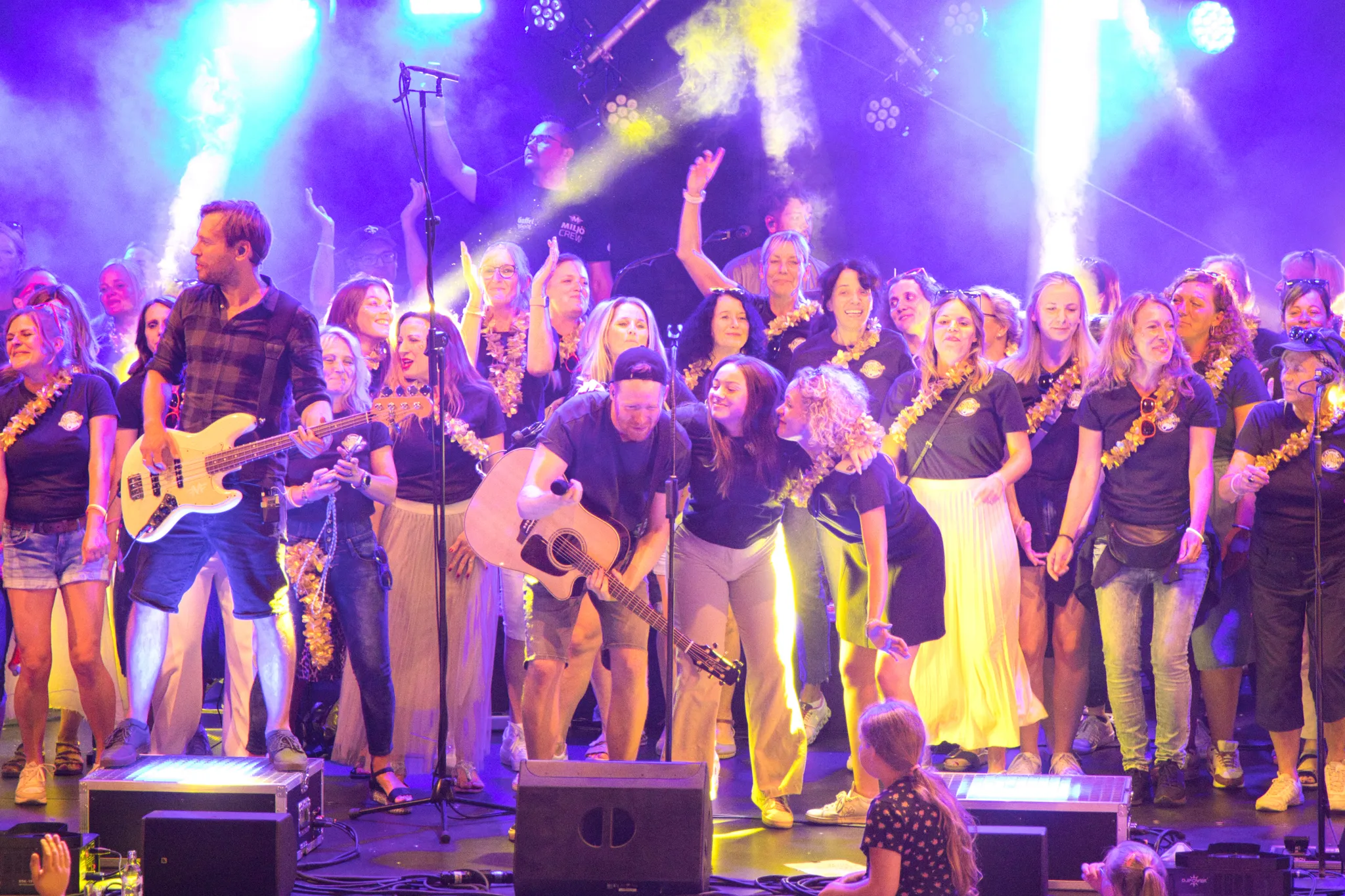 Weiter Blick auf die nebelverhangene Bühne mit Chor und Band in grün-violettem Scheinwerferlicht, Publikum hält leuchtende Handys in die Höhe.