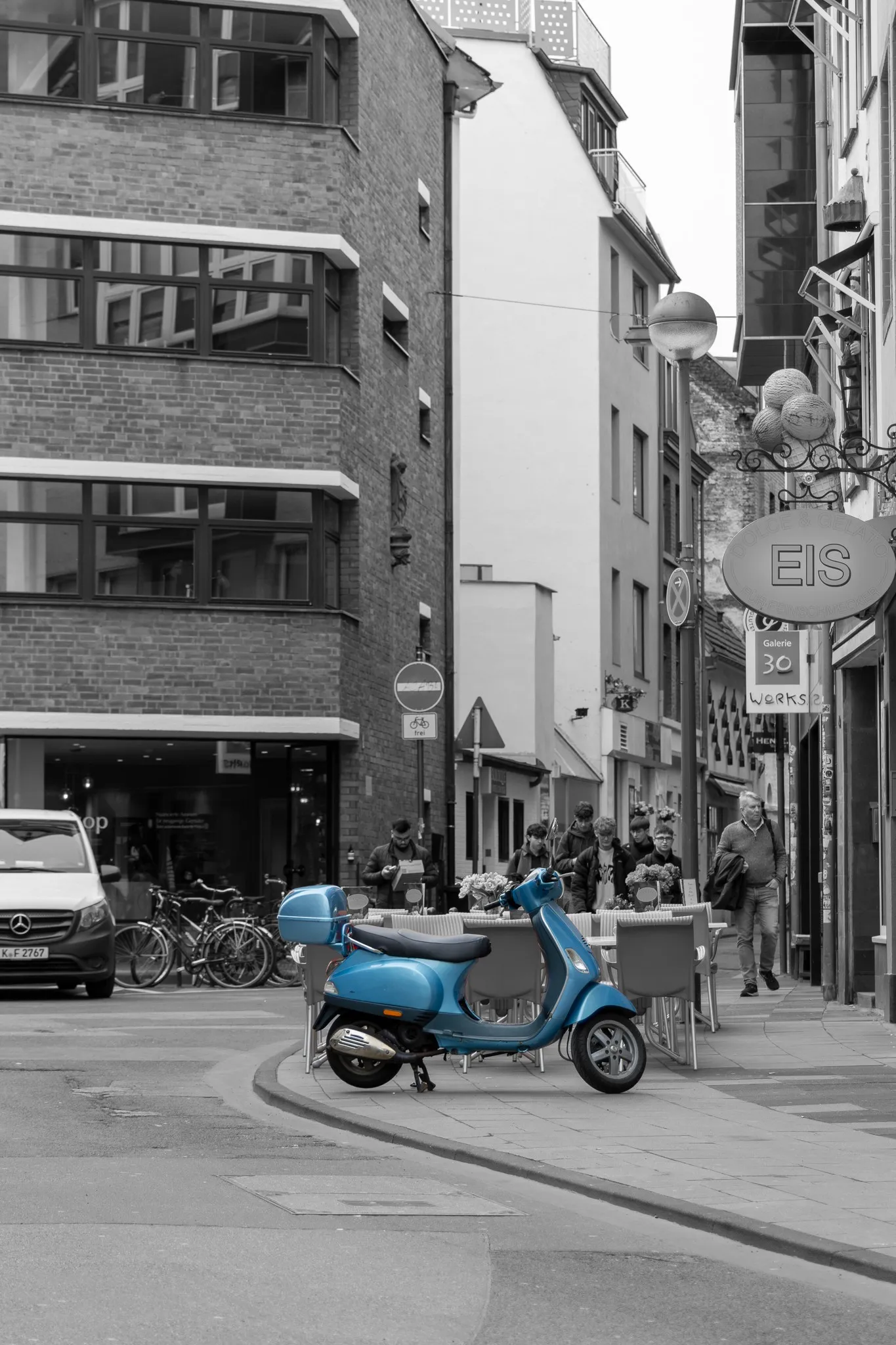 Blauer Motorroller auf einer Kopfsteinpflaster-Straßenecke, freigestellt in Farbe vor einer sonst schwarz-weißen Altstadtszene mit Cafégästen und Fahrrädern im Hintergrund.