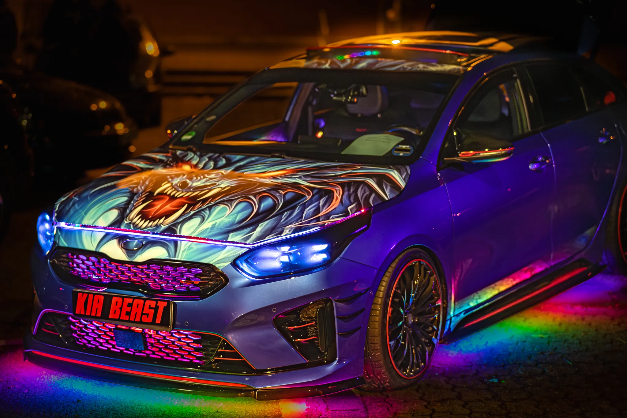 Blauer Kompaktwagen mit aufwendiger Drachen-Airbrush-Lackierung und mehrfarbiger Unterbodenbeleuchtung bei Nacht, Kennzeichenhalter mit Schriftzug „Kia Beast“.